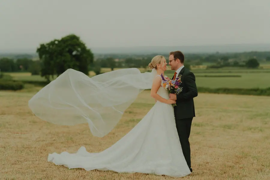 Alice and Sean in the field with her veil lifted by the wind