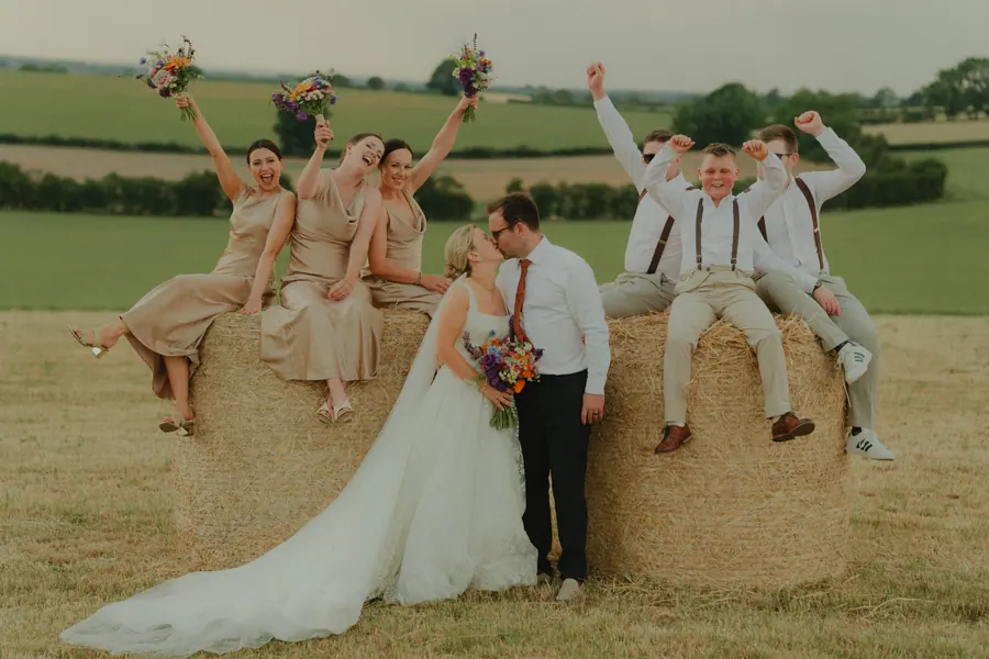 The wedding party raising bouquets and arms around a round hay bale