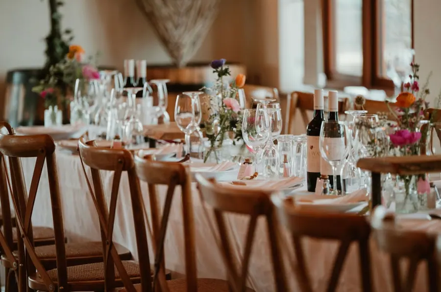 Long wooden dinner table laid with flowers, bottles and brown chairs