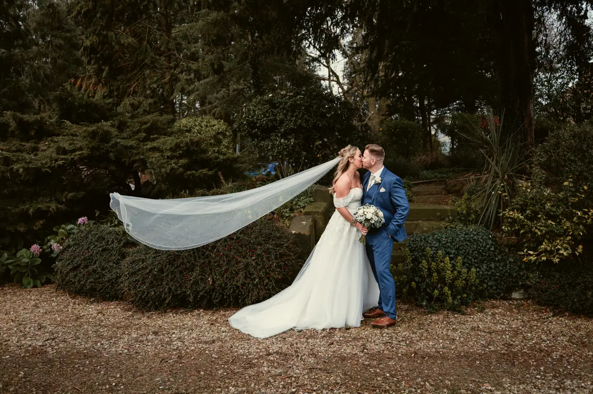 The couple embracing on gravel with Hayley's veil spread behind them