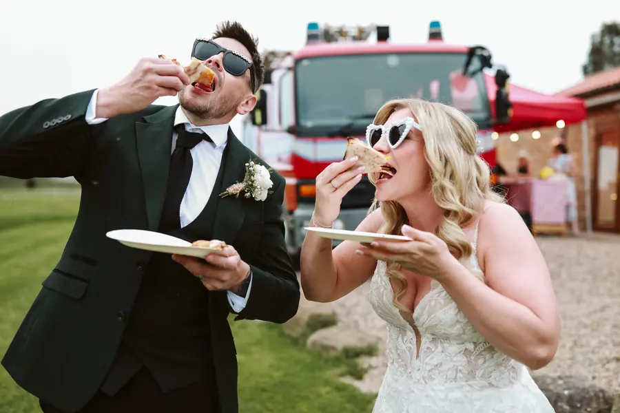 Lauren and Iain eating pizza beside a red food truck in white sunglasses