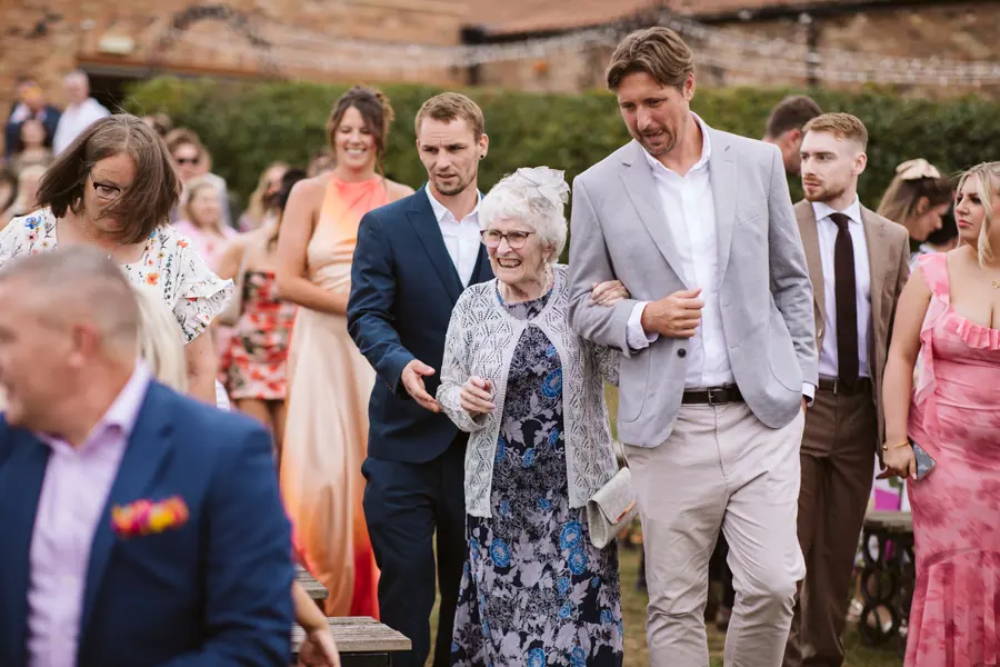A guest accompanying an older woman along the outdoor ceremony aisle