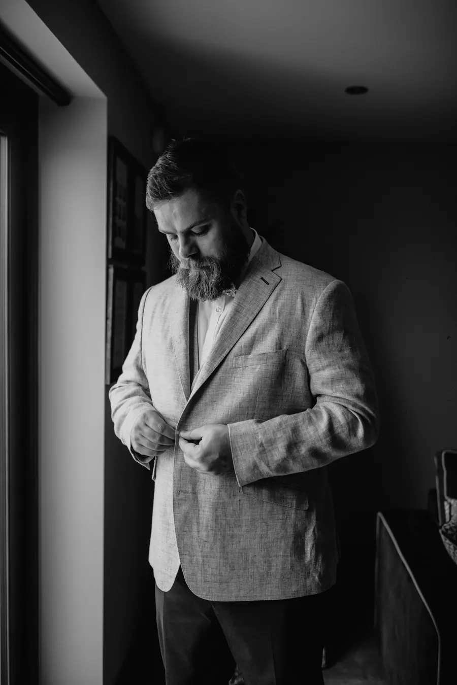 Martin fastening his pale jacket beside a dark window in black and white