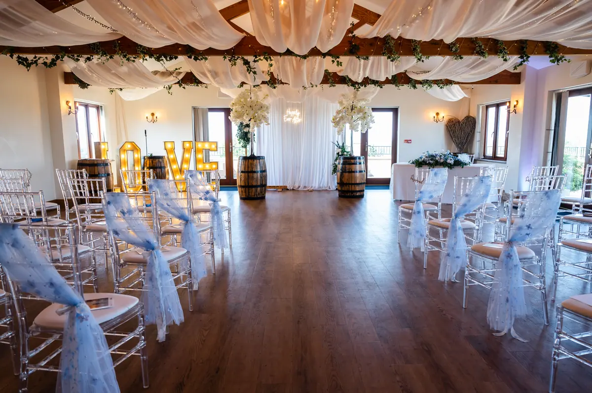 Clear ceremony chairs facing a greenery-wrapped wooden arch in the Hall