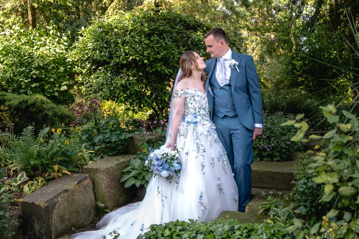 Megan and Dan standing among leafy plants beside a low stone wall