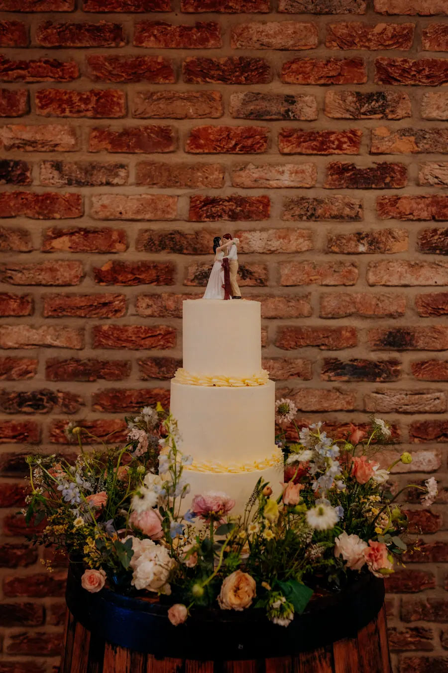 Pale tiered cake surrounded by flowers against a red brick wall