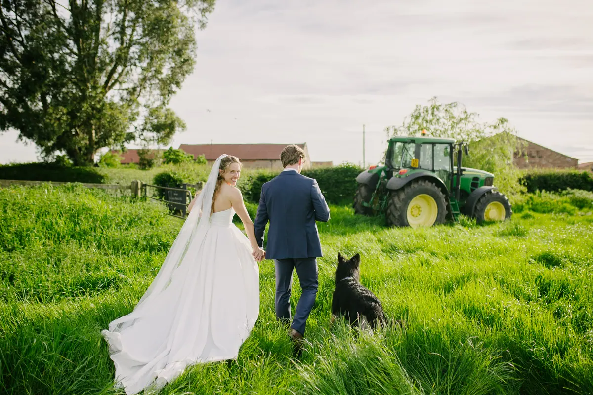 Rachel and Andrew walking through grass with a black dog near a tractor