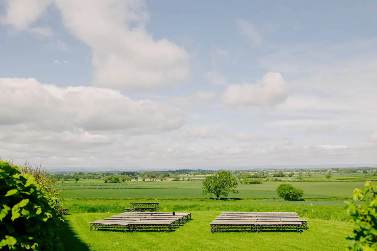 Wooden ceremony benches facing wide green fields beneath broken cloud