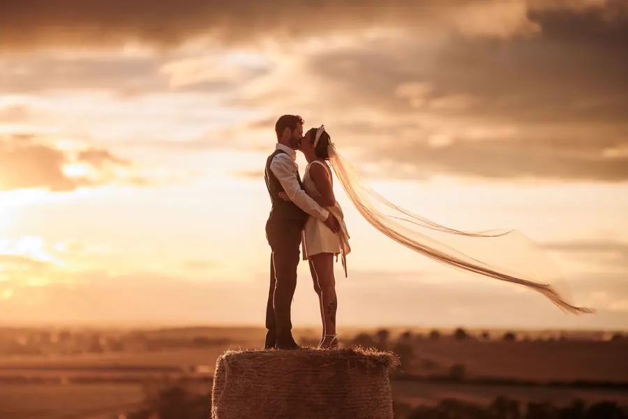 Sophie and Ben standing on a hay bale as her veil crosses the sunset
