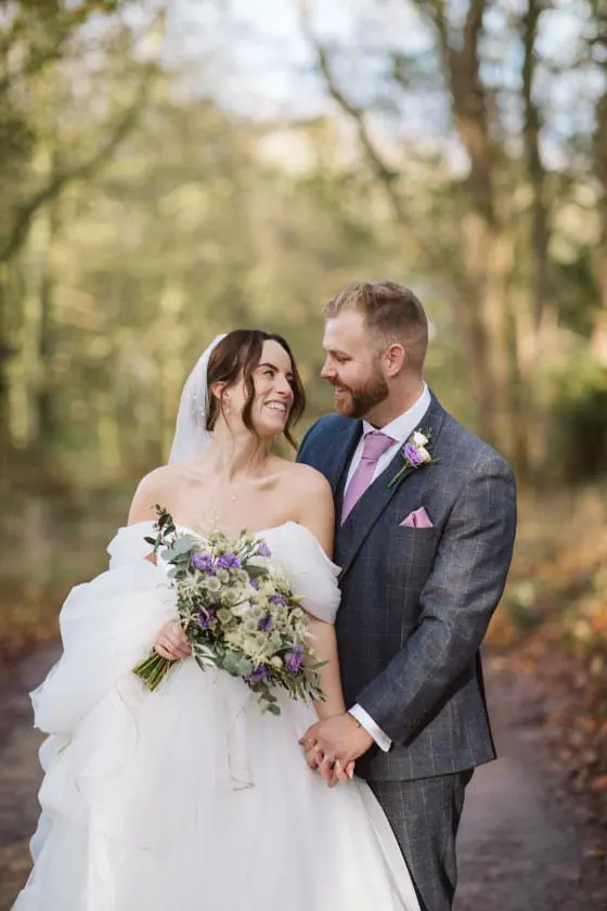 The couple holding hands beside woodland with purple flowers between them