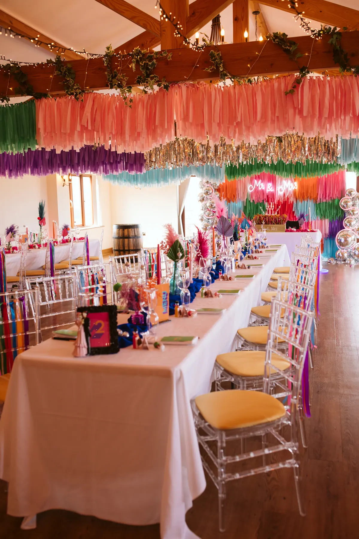 Colourful streamers above dressed wedding tables and clear chairs beneath Thief Hall’s beams