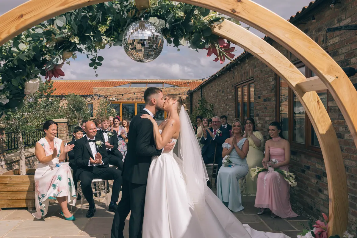 Newlyweds kissing beneath a flower-covered wooden arch and mirror ball in the Thief Hall courtyard